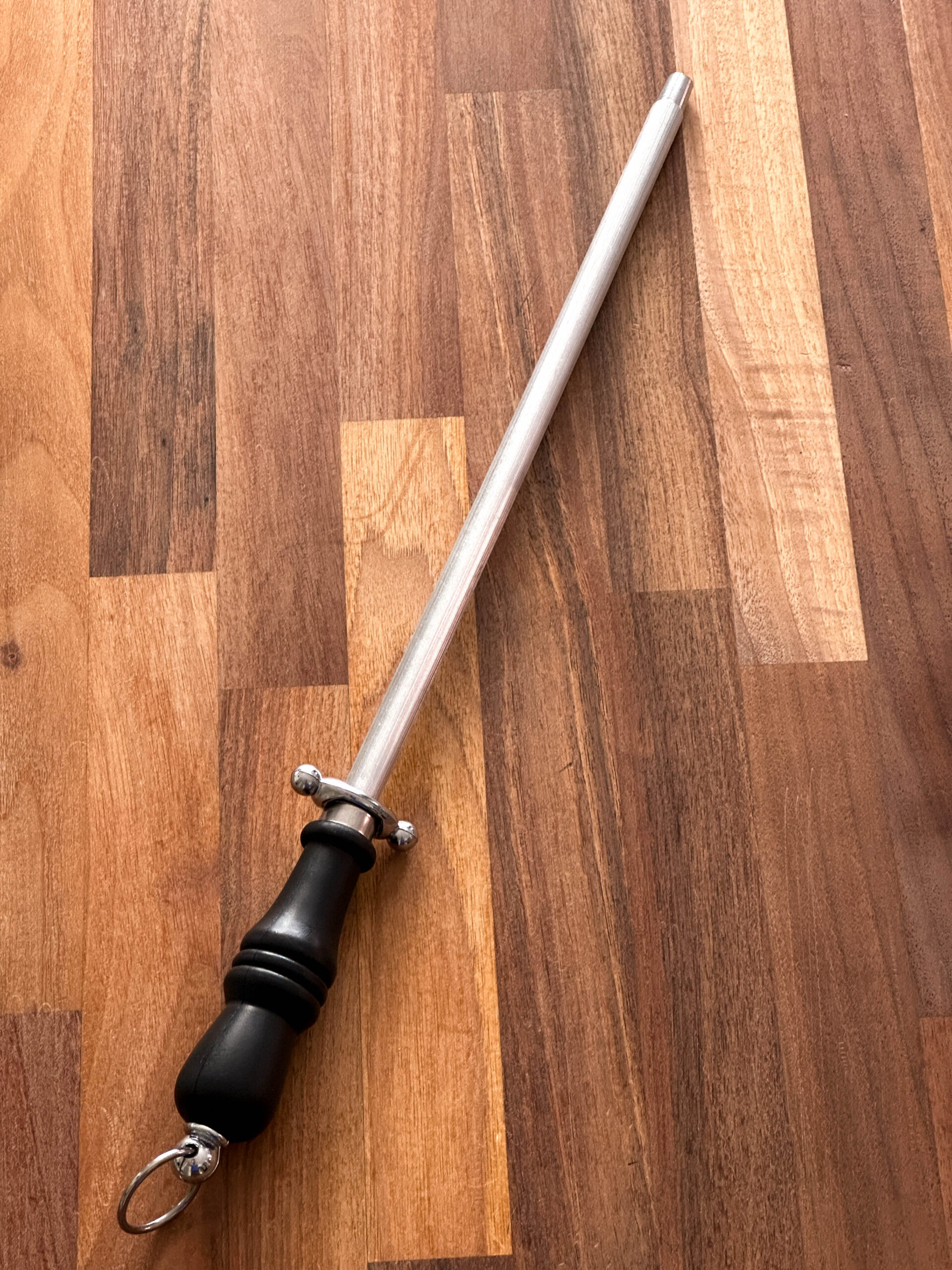 Metal honing steel for knives, with black plastic handle and silver hanging loop. Pictured against a wooden background