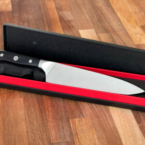 Metal chef knife with black handle, in a red and black presentation box. Pictured on a wooden background