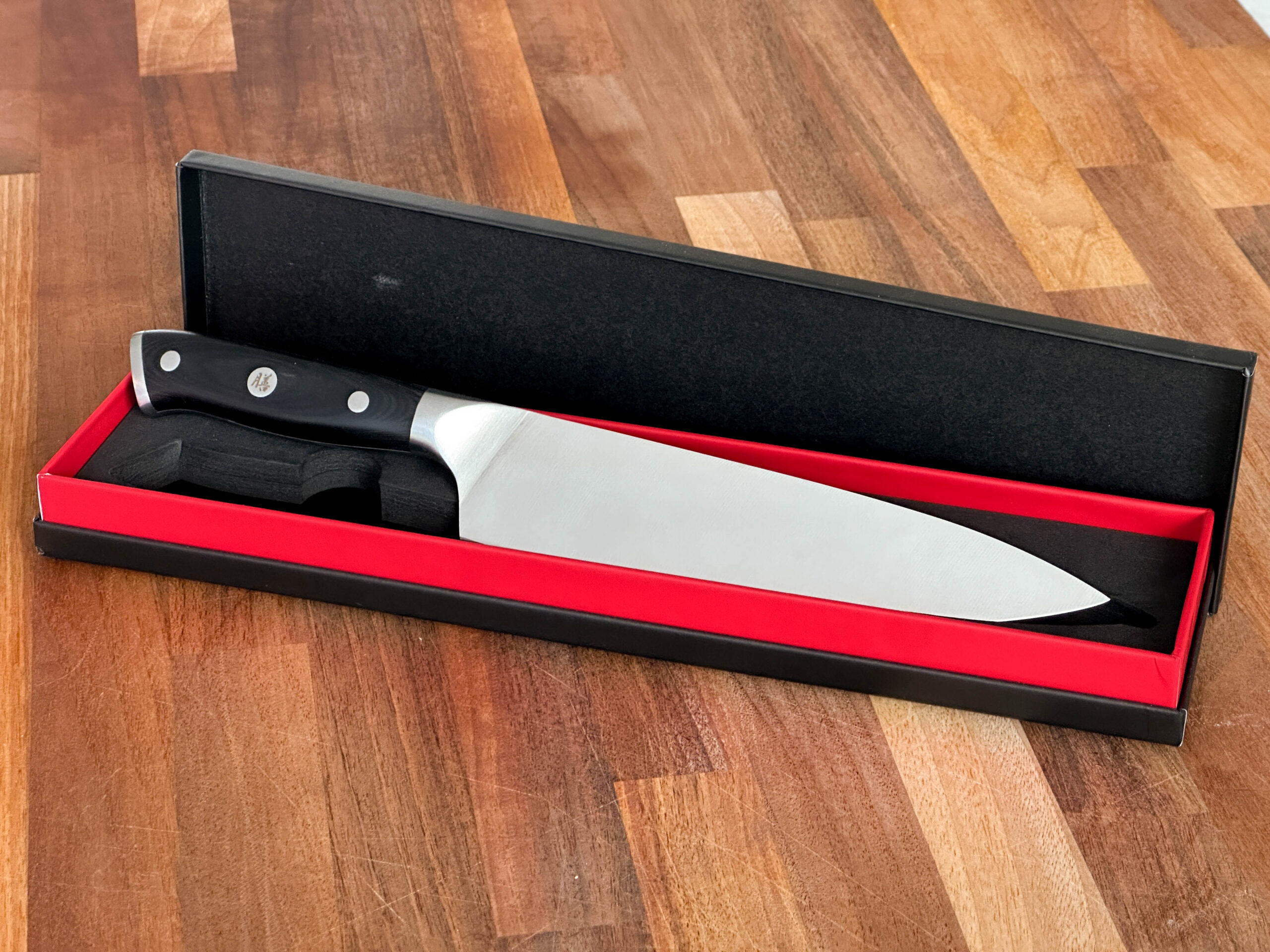 Metal chef knife with black handle, in a red and black presentation box. Pictured on a wooden background